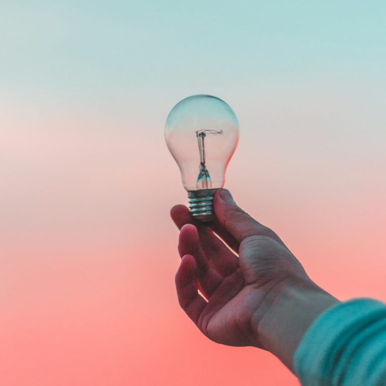 Eine Hand hält eine Glühbirne vor einem pastellfarbenen Hintergrund.