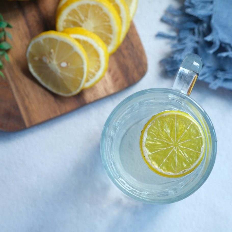 Ein Glas Wasser mit einer Zitronenscheibe, daneben geschnittene Zitronen auf einem Holzbrett.