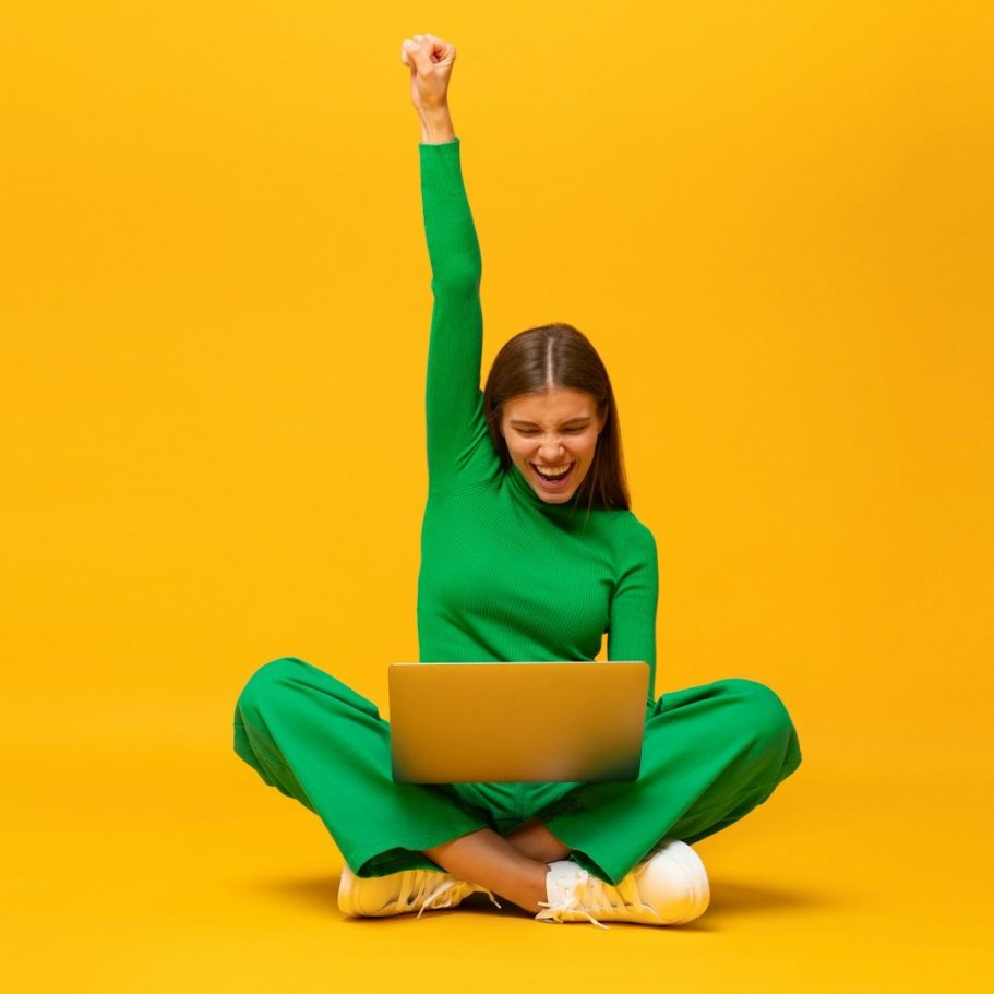 Junge Frau in grüner Kleidung sitzt mit Laptop und hebt begeistert die Faust in die Luft.