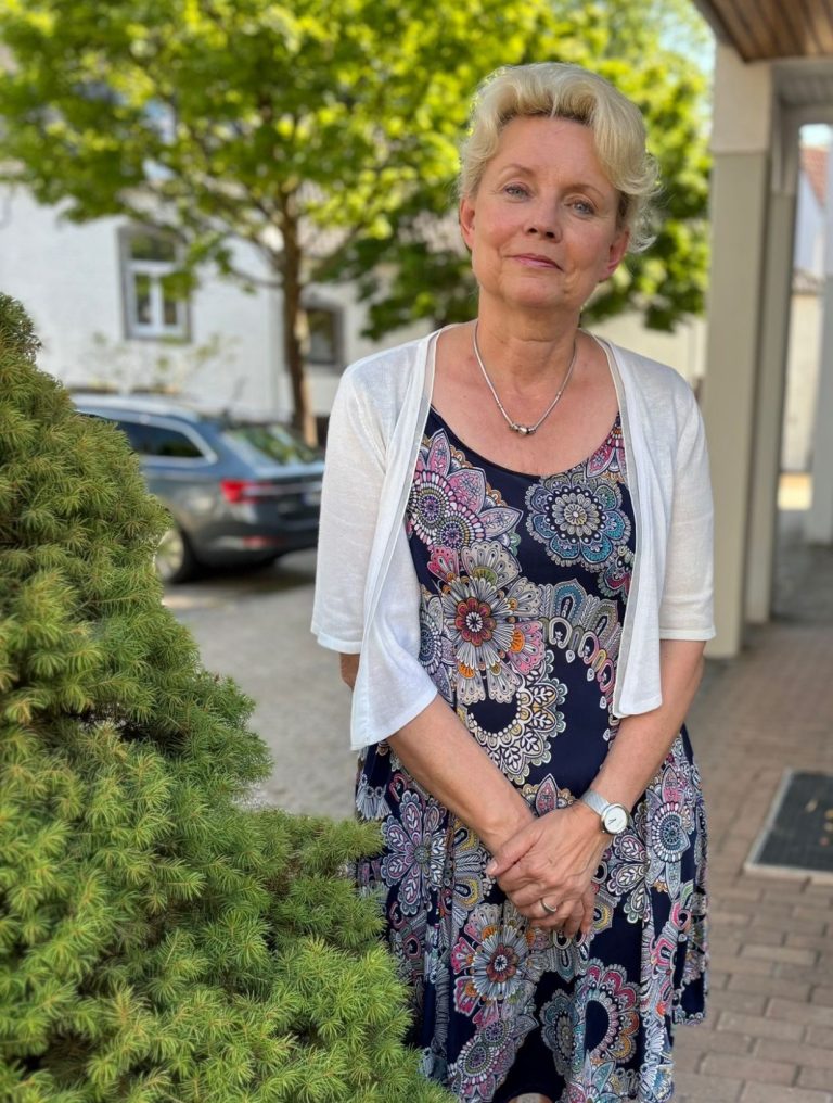 Ulla Malejka, Standortleitung Laurensberg Frau mit blonden Haaren in einem bunten Kleid steht neben einem Baum im Freien.
