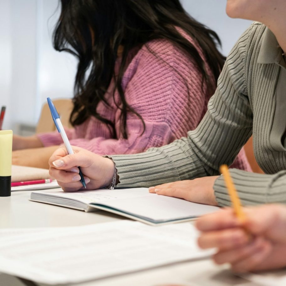 Hände von Personen, die in Notizbücher schreiben, mit Stiften und einem Kalender auf dem Tisch.