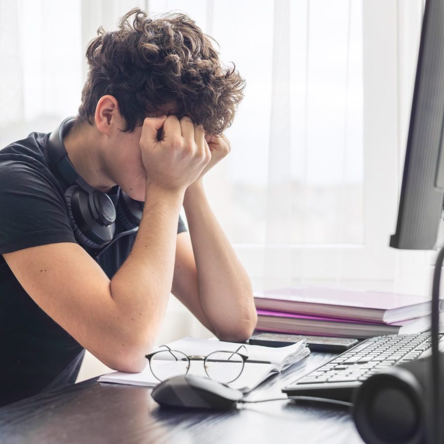 Junger Mann sitzt am Schreibtisch und hält sich den Kopf, sichtbar gestresst.