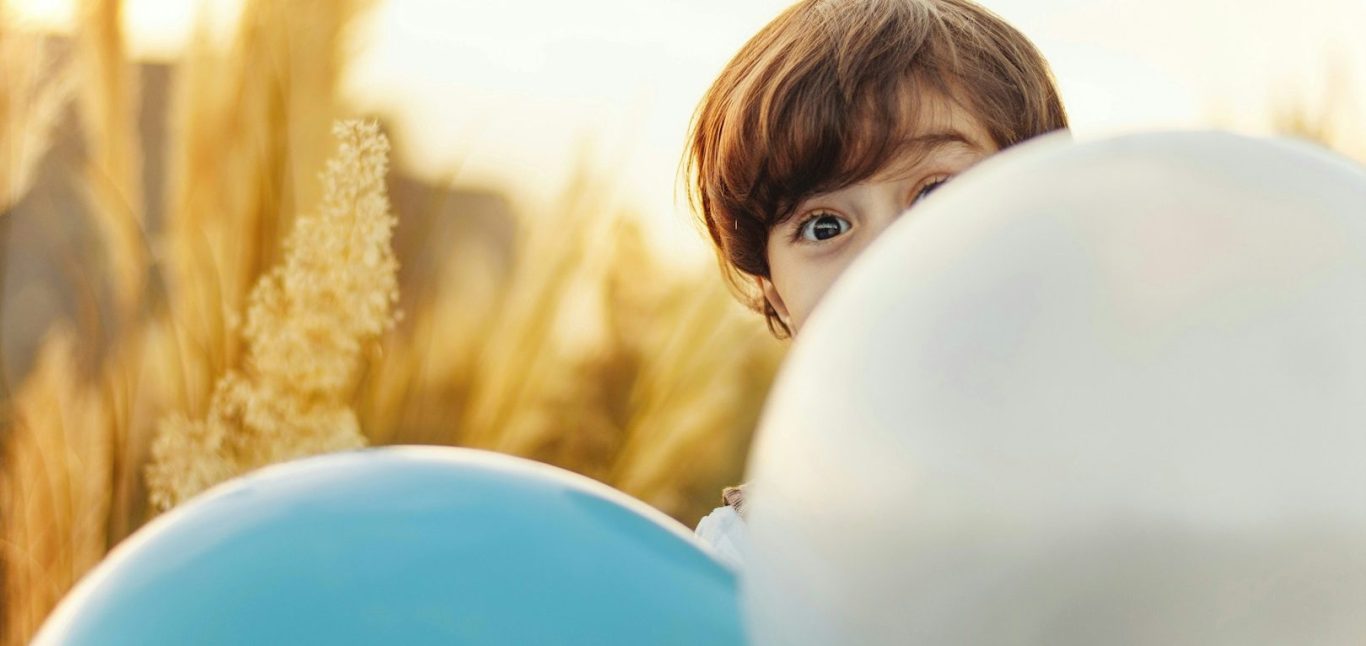 Neugieriger Junge hinter Luftballons Ein Junge schaut neugierig hinter bunten Luftballons hervor in einem Feld.