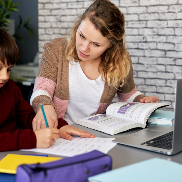 Eine Lehrerin hilft einem Jungen beim Lernen am Tisch mit Büchern und einem Laptop.