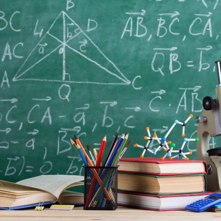 Mathe- und Chemie-Utensilien auf einem Tisch vor einer Tafel mit Formeln.