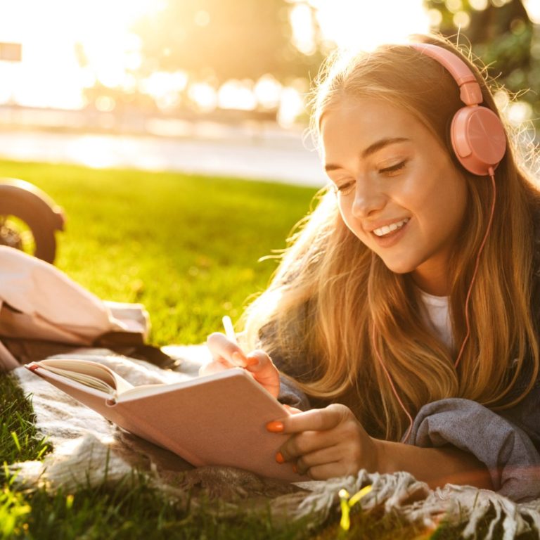 Junge Frau mit Kopfhörern, die auf einer Wiese in ein Notizbuch schreibt.
