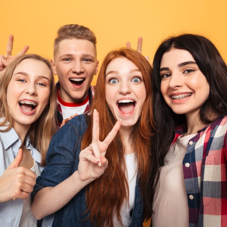 Vier junge Menschen lächeln und zeigen Frieden-Zeichen vor gelbem Hintergrund.