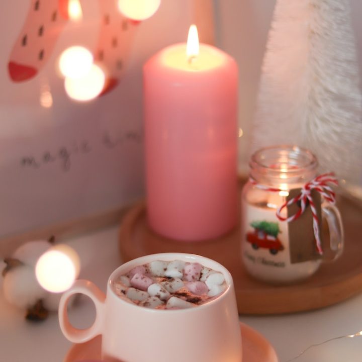 Rosefarbene Kerze im Hintergrund, vorne Tasse mit heißer Schokolade, Marshmallows und Weihnachtsdeko auf einem Tisch.
