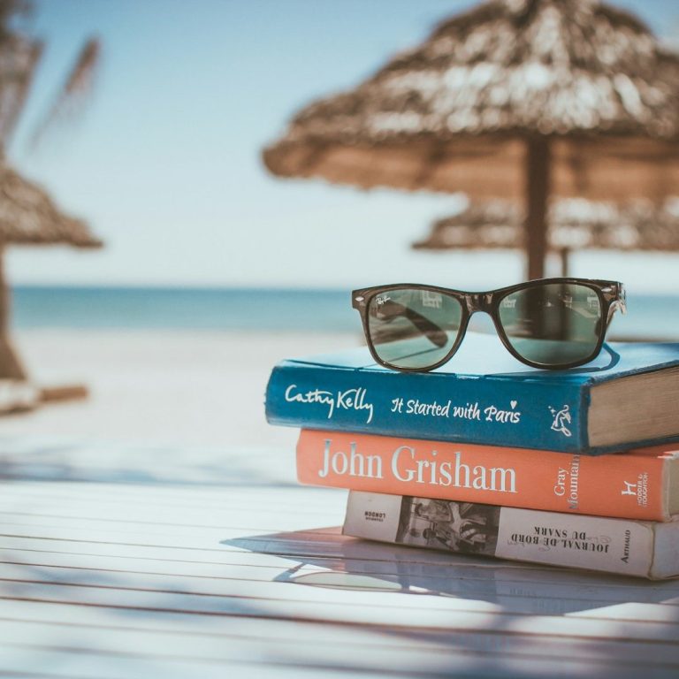 Lesen und Lernen in den Ferien Sonnenbrille auf gestapelten Büchern am Strand mit Palmen und Meer im Hintergrund.