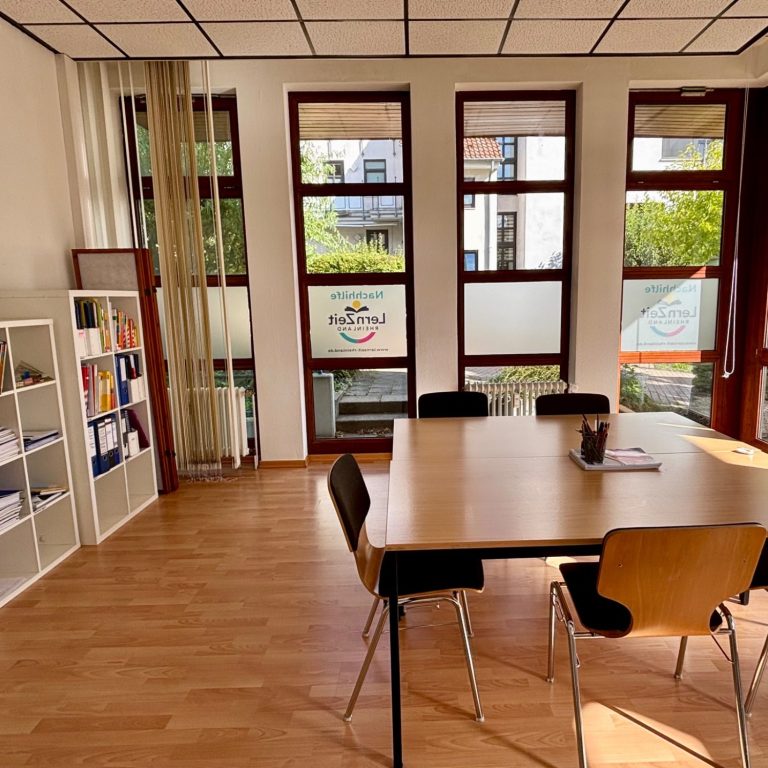 Heller Unterrichtsraum mit Tisch, Stühlen und Regalen, großen Fenster mit Blick ins Freie.